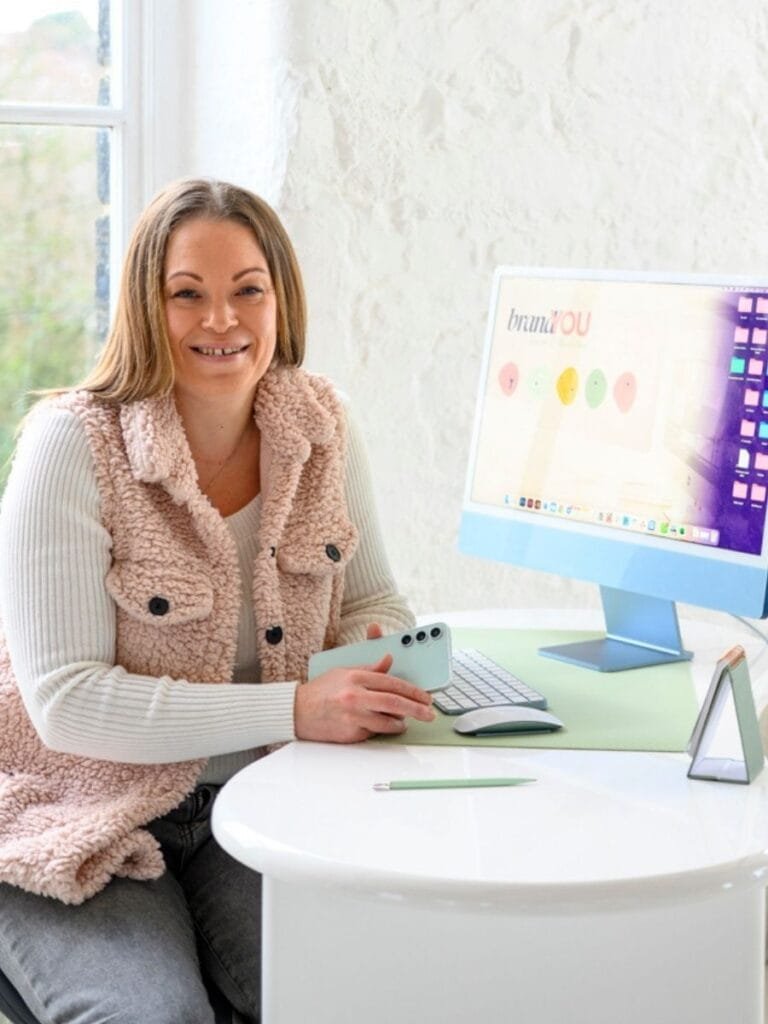 Founder of Brand You Design & Marketing sitting at a desk with a computer, phone and brand visuals on screen